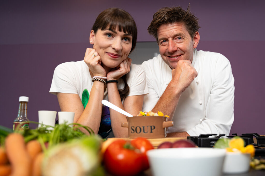 Kristina vom Dorf und Jörg Färber lehnen an einem Tisch mit verschiedenen Gemüsen und Gewürzen und einem Topf mit der Aufschrift Soup.