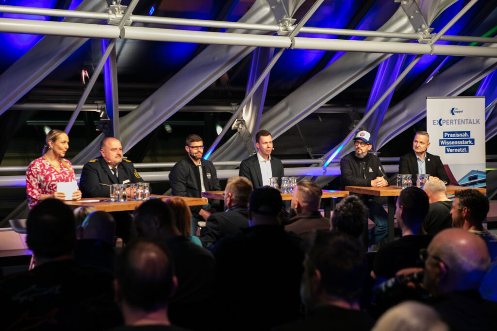 Eine Frau und sechs Männer sitzen in einem verklasten Raum in Abendstimmung auf einer kleinen Bühnen an einem Tisch. Im Vordergrund sitzen Menschen und hören zu