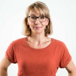 Bild einer Frau mit blonden, mittellangen Haare und Brille mit dunklem Rand. Sie trägt ein orangnes T-Shirt und schaut freundlich.